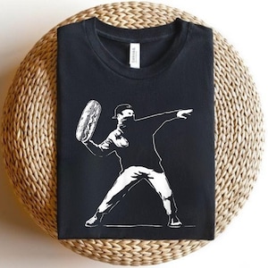 May include: Black t-shirt featuring a white graphic of a person throwing a hot dog. The design is a stylized illustration, with the figure wearing a cap and a face covering. The shirt is displayed on a woven surface.