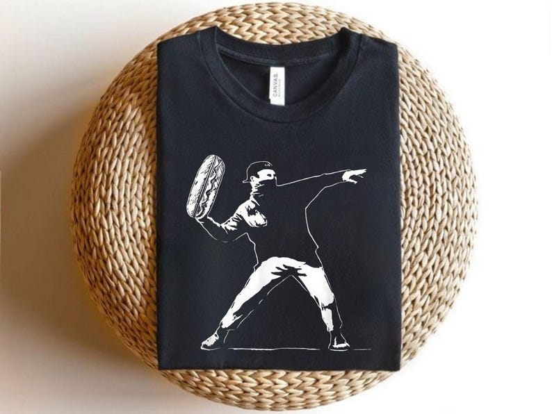 May include: Black t-shirt featuring a white graphic of a person throwing a hot dog. The design is a stylized illustration, with the figure wearing a cap and a face covering. The shirt is displayed on a woven surface.