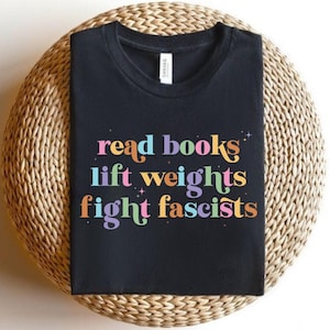 Op de afbeelding: Zwart t-shirt met de tekst "read books lift weights fight fascists" in kleurrijke, retro letters. Het shirt ligt op een ronde, geweven ondergrond, wat suggereert dat het geschikt is voor dagelijks gebruik.
