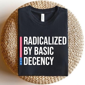 Op de afbeelding: Zwart T-shirt met de tekst "RADICALIZED BY BASIC DECENCY" in het wit. Een verticale streep met rode en witte strepen en blauwe sterren staat links van de tekst. Het shirt ligt op een geweven, cirkelvormig oppervlak.