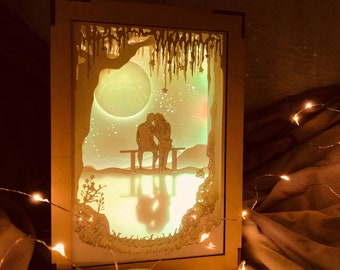 May include: A wooden shadow box featuring a laser-cut design of a couple seated on a bench beneath a crescent moon and stars. The box is illuminated from behind, casting a warm glow.