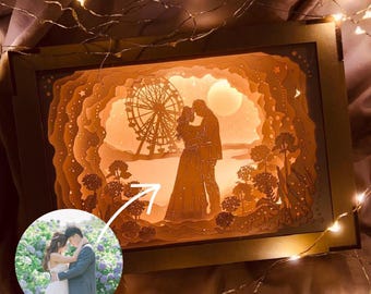以下が含まれることがあります： カップルがシルエットで描かれた3Dレーザーカットの木製シャドウボックス。シーンには観覧車、月、星空が描かれています。カップルは湖の前に立っています。左下隅にはカップルの小さな写真があります。
