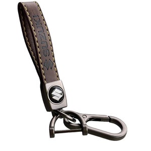 May include: Brown leather keychain with a metal loop and a silver charm featuring the Suzuki logo. The leather strap has yellow stitching and the word "SUZUKI" embossed on it.