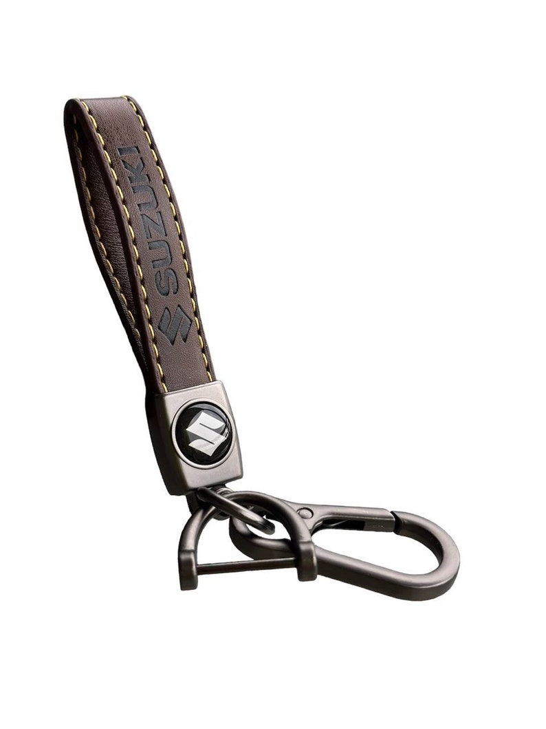 May include: Brown leather keychain with a metal loop and a silver charm featuring the Suzuki logo. The leather strap has yellow stitching and the word "SUZUKI" embossed on it.