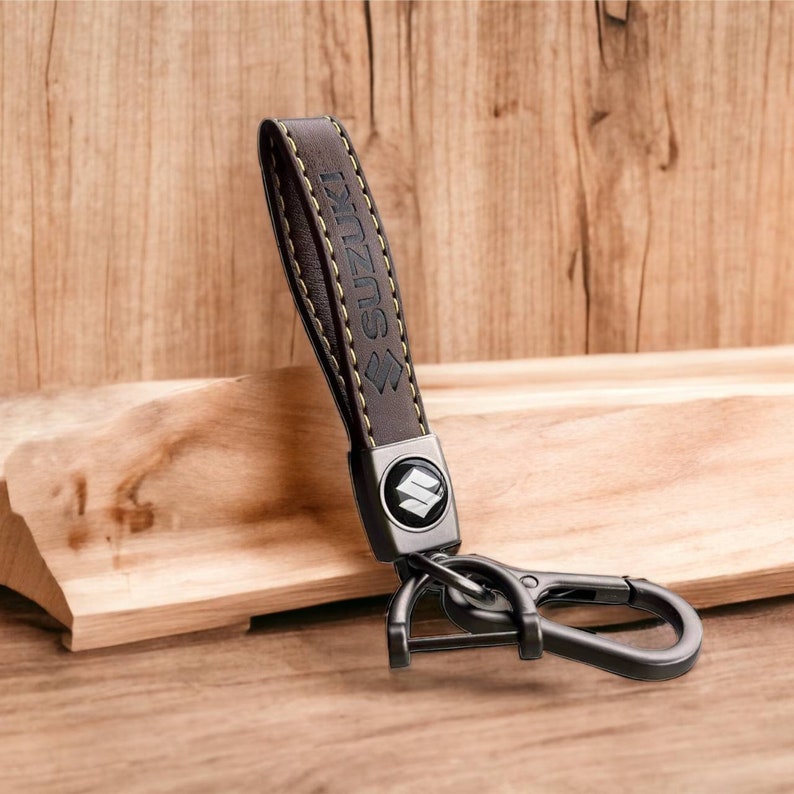 May include: A brown leather keychain with a metal Suzuki logo and a silver key ring. The keychain has yellow stitching.