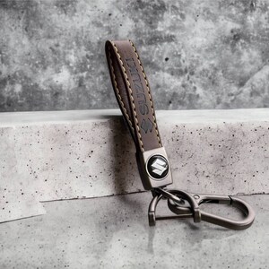 May include: Brown leather keychain with a metal Suzuki logo and a silver hook. The keychain has yellow stitching.