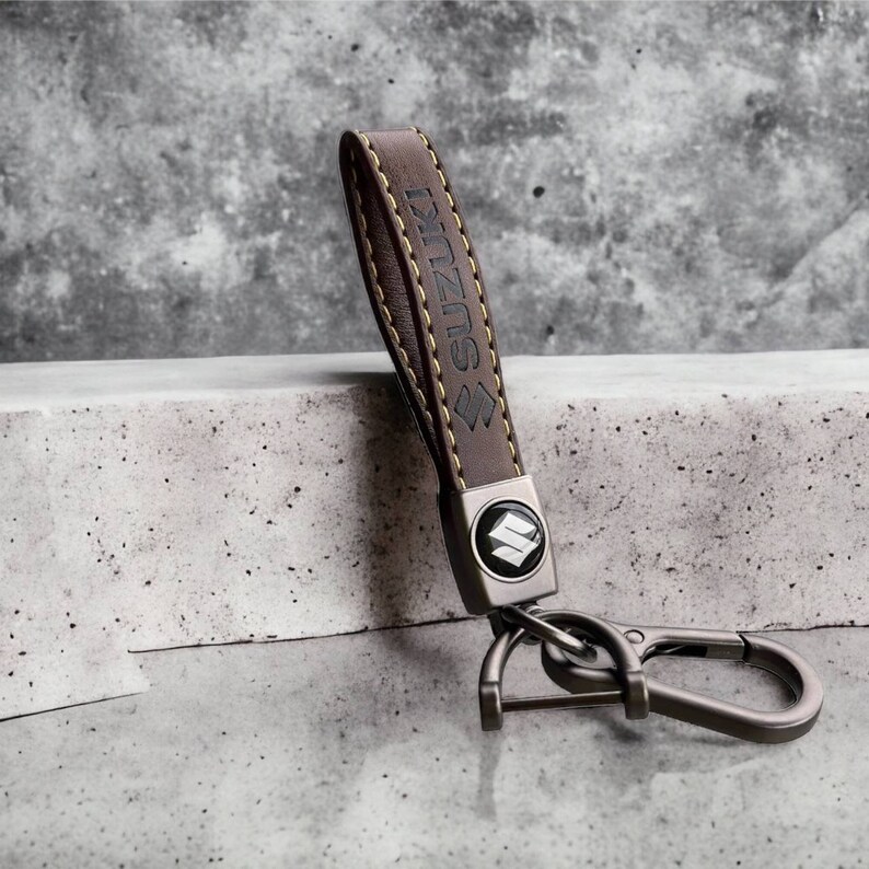 May include: Brown leather keychain with a metal Suzuki logo and a silver hook. The keychain has yellow stitching.