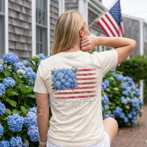 May include: Cream-colored t-shirt featuring a floral American flag design on the back. The flag's stars are replaced with blue hydrangea flowers, and the stripes are red. The text "Sweet Land of Liberty" is printed below the flag.