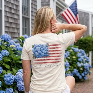 May include: Cream-colored t-shirt featuring a floral American flag design. The flag incorporates blue hydrangea flowers in the upper left corner and red stripes. The text "Sweet Land of Liberty" and "Est. 1776" are printed below the flag.