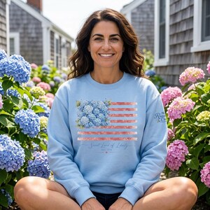 Peut inclure: Sweat-shirt bleu clair orné d'un drapeau américain floral. Le drapeau incorpore des fleurs d'hortensia bleues et des rayures rouges, avec le texte "Sweet Land of Liberty" et "Est. 1776". Le logo Rogue Crew est sur la manche droite.