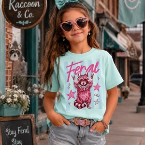 以下が含まれることがあります： 水色のTシャツを着た若い人がいます。ピンク色の「Feral」という文字とアライグマのグラフィックがプリントされています。デニムのショートパンツ、ピンクのハート型のサングラス、ヘアリボンも着用しています。Tシャツはストライプの背景です。