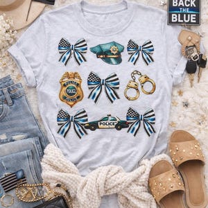 以下が含まれることがあります： 警察をテーマにしたグラフィックが特徴のライトグレーのTシャツ。リボン、警察バッジ、パトカー、手錠などが描かれています。シャツには警察の帽子のデザインもあります。アクセサリーには、黒いハンドバッグ、サンダル、細い青い線の旗が含まれています。