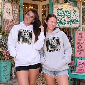 May include: A white hooded sweatshirt and a light grey hooded sweatshirt, both featuring a black and white portrait of a woman with the text "GUTS, GRIT & LIPSTICK" printed in black letters inside a gold-toned rectangular frame on the chest.