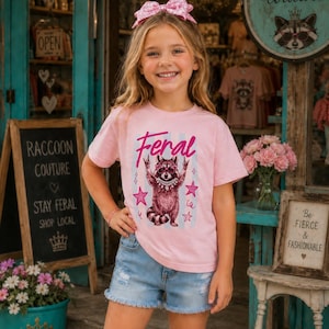 以下が含まれることがあります： 「Feral」の文字とアライグマのグラフィックがプリントされた、明るいピンク色のTシャツを着た若い人。アライグマはサングラスとネックレスを着用。デニムのショートパンツとピンクのリボンも着用しています。