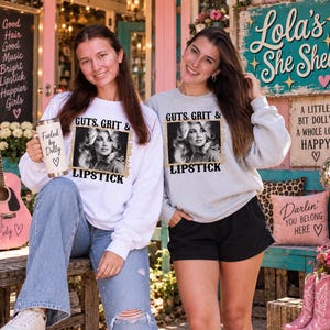 May include: A white long-sleeve sweatshirt and a grey long-sleeve sweatshirt, both featuring a black and white portrait of a woman with the text GUTS, GRIT & LIPSTICK. The white sweatshirt is paired with a tumbler labelled Fueled by Dolly.