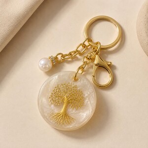 Op de afbeelding: Een goudkleurige sleutelhanger met een ronde, parelmoerwitte hars hanger met een ingebed gouden levensboomontwerp. De sleutelhanger bevat een gouden splitring, een karabijnsluiting en een korte ketting met een enkele witte parelkraal.