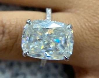 May include: A large, cushion-cut diamond engagement ring set in a white gold band with a pave setting. The ring is on a person's finger.
