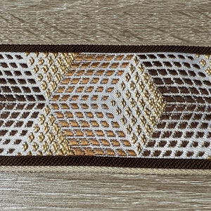 Könnte beinhalten: Nahaufnahme eines dekorativen gewebten Bandes mit geometrischem Muster. Das Band weist ein sich wiederholendes Design aus Rauten und Zickzacklinien in Braun-, Beige- und Weißtönen auf. Die Ränder sind mit einer dunkelbraunen Bordüre eingefasst.