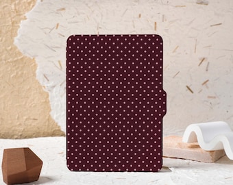 May include: A burgundy case with a repeating pattern of small, light pink polka dots. The case has a rectangular shape with rounded corners and a side flap. The background is a textured, off-white surface.