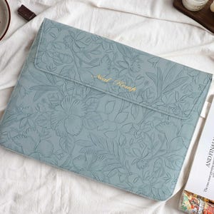 Op de afbeelding: Een lichtblauwe laptoptas met een bloemenpatroon en de gouden tekst "Neal Kemp". De tas is afgesloten met een flap. Rechts ligt een tijdschrift met de tekst "AND FINALLY..."