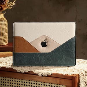 Puede incluir: Una funda para portátil multicolor con un diseño de montaña. La funda presenta una parte superior blanca, una sección media marrón y una parte inferior verde oscuro. Se ve un recorte del logotipo de Apple. La funda está sobre una mesa de madera con un tapete de encaje.