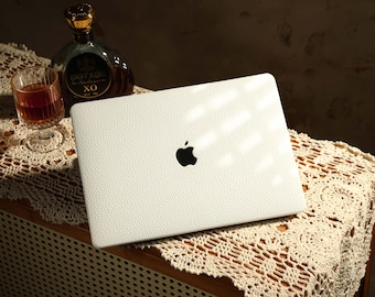 Op de afbeelding: Een witte laptop met een zwart Apple-logo op de deksel. De laptop staat op een wit gehaakt tafellaken op een houten oppervlak.