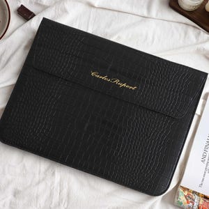 Puede incluir: Funda negra para portátil de piel sintética con un patrón de cocodrilo texturizado. La solapa tiene el texto dorado "Carlos Rupert". La funda está sobre una superficie blanca con chocolate y una revista.
