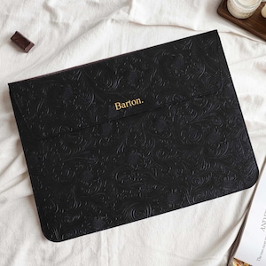 Könnte beinhalten: Schwarze Laptoptasche mit geprägtem floralem Muster. Der Text "Barton" ist in Goldbuchstaben gehalten. Die Hülle ist rechteckig mit einem Klappenverschluss. Schokoladenriegel und eine Zeitschrift sind im Hintergrund.