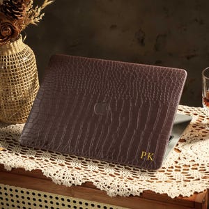 May include: A brown laptop case with a crocodile skin texture, featuring the initials "PK" in gold. The case is on a laptop, resting on a cream-colored lace tablecloth. A glass of amber liquid sits nearby.