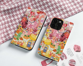 May include: Two phone cases featuring vibrant illustrations of fruits, jars, and beverages. They rest on a white surface, with a pink and white patterned fabric in the background. A sticker with the text "LIFE IS GOOD" is also visible.