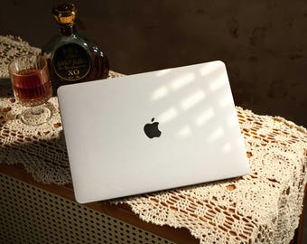Op de afbeelding: Een zilveren laptop met het Apple-logo op de deksel. De laptop staat op een wit gehaakt tafellaken op een houten oppervlak.