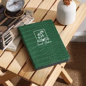 Peut inclure: Un journal vert en similicuir avec une texture crocodile, une fermeture éclair noire et le nom "Sarah Baber" imprimé sur la couverture. La couverture présente un motif floral dans un cadre rectangulaire. Le journal est sur une table d'appoint en bois.