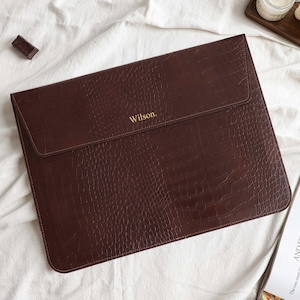 May include: A brown leather laptop sleeve with a crocodile texture. The word "Wilson" is embossed in gold on the front. The sleeve is closed with a flap. Chocolate bars and a magazine are in the background.