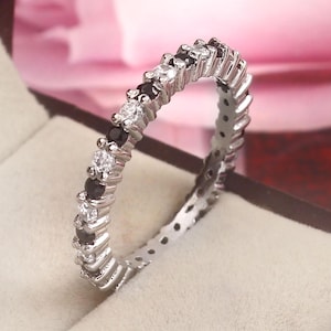 May include: A silver ring featuring alternating black and clear gemstones. The ring's design is a zig-zag pattern, with the gemstones evenly spaced around the band. The ring is presented in a jewellery box against a pink backdrop.