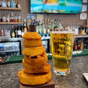 May include: A stack of golden-brown onion rings on a small wooden base, topped with a small wooden cylinder. A tall glass of beer sits next to the onion rings on a bar. The background shows a bar with bottles and a television.