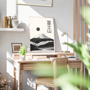 May include: A framed print of Mount Fuji with Japanese characters, a black sun, and a black and white mountain landscape. The print is on a wooden desk with a small plant, books, and a gold desk lamp. A wooden chair is in front of the desk.