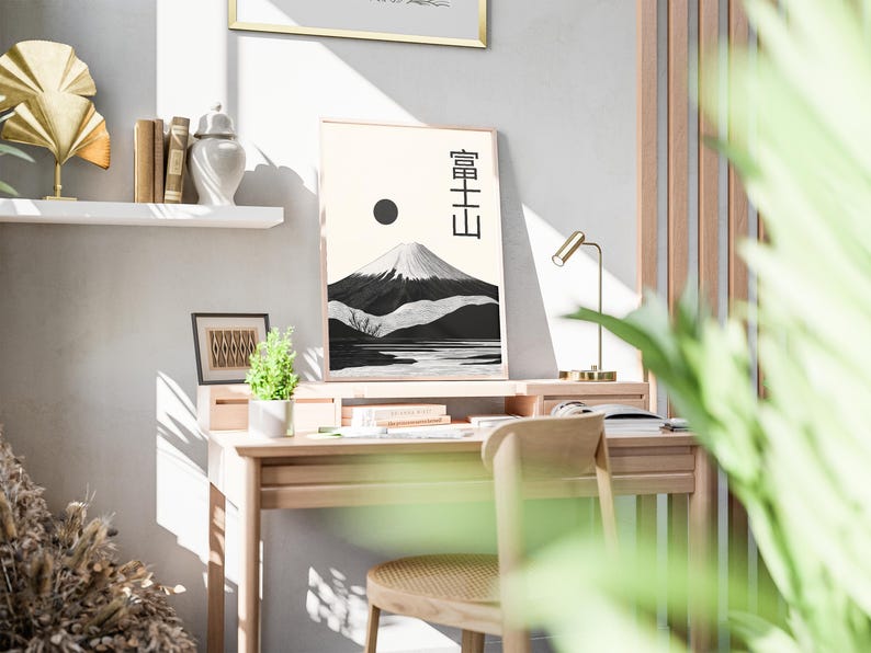 May include: A framed print of Mount Fuji with Japanese characters, a black sun, and a black and white mountain landscape. The print is on a wooden desk with a small plant, books, and a gold desk lamp. A wooden chair is in front of the desk.