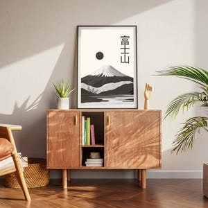 May include: A framed black and white art print featuring a mountain landscape with Japanese text. The artwork is displayed on a wooden cabinet with a plant and a wooden hand sculpture. The room has a modern, minimalist aesthetic.