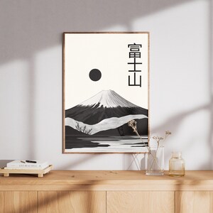 May include: A framed art print featuring a black and white illustration of Mount Fuji with a snow-capped peak, a black sun, and Japanese text. The artwork is displayed on a light-colored wall, with a wooden cabinet in the foreground.