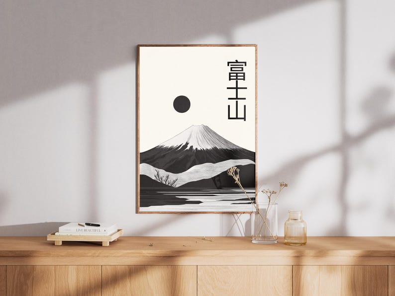 May include: A framed art print featuring a black and white illustration of Mount Fuji with a snow-capped peak, a black sun, and Japanese text. The artwork is displayed on a light-colored wall, with a wooden cabinet in the foreground.