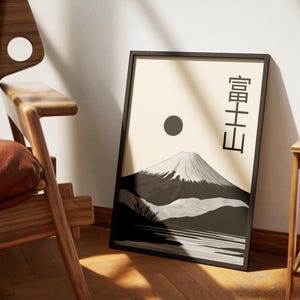May include: Framed art print featuring a black and white illustration of Mount Fuji with a sun and Japanese text. The artwork is displayed on a light beige background. The print is in a black frame and leans against a white wall.