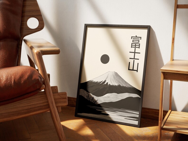 May include: Framed art print featuring a black and white illustration of Mount Fuji with a sun and Japanese text. The artwork is displayed on a light beige background. The print is in a black frame and leans against a white wall.