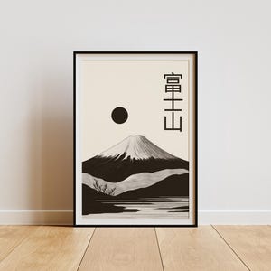 May include: Black framed art print featuring a minimalist illustration of Mount Fuji. The artwork depicts the mountain with a snow-capped peak, a dark sun, and Japanese characters. The print is displayed on a wooden floor.