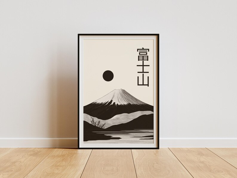 May include: Black framed art print featuring a minimalist illustration of Mount Fuji. The artwork depicts the mountain with a snow-capped peak, a dark sun, and Japanese characters. The print is displayed on a wooden floor.