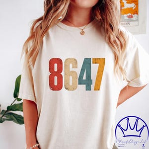 Può includere: T-shirt color crema con il numero "8647" stampato in colori retrò: rosso, beige, verde acqua e giallo. La maglietta ha una vestibilità ampia e maniche corte. Il design è centrato sul davanti.