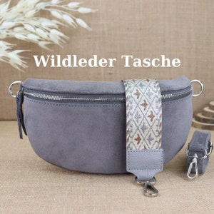 Può includere: Una marsupio in pelle scamosciata grigia con una cerniera argentata e una tracolla a fantasia. La tracolla è realizzata in tessuto beige e bianco con un motivo geometrico. Il testo "Wildleder Tasche" è scritto in bianco su uno sfondo marrone.