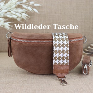 Può includere: Una borsa a tracolla in pelle scamosciata marrone con una tracolla a motivo bianco e marrone. La borsa ha una chiusura a zip e una fibbia argentata. Il testo "Wildleder Tasche" è scritto in bianco su sfondo marrone.