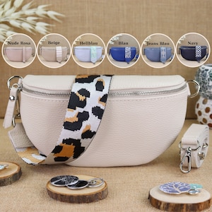 Può includere: Una marsupio in pelle bianca con chiusura a zip e una tracolla regolabile con stampa leopardata. La borsa è mostrata su una superficie marrone con una varietà di piccoli oggetti decorativi.