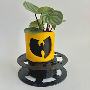 May include: A yellow and black planter with a Wu-Tang Clan logo, holding a small plant with green and silver striped leaves. The planter sits on a black spool-like stand, creating a unique and modern aesthetic. The plant has red stems.