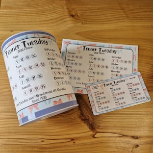 May include: A collection of "Tenner Tuesday" calendars, featuring a cylindrical container and two cards. The design is light blue and white, with bow accents and calendar dates. The text "2026 Edition" is displayed on the items.